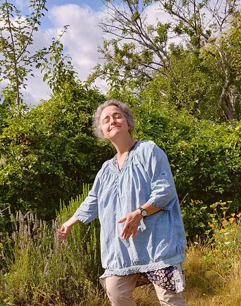 Anne, yeux fermés sous le soleil, mains effleurant la végétation d'un jardin